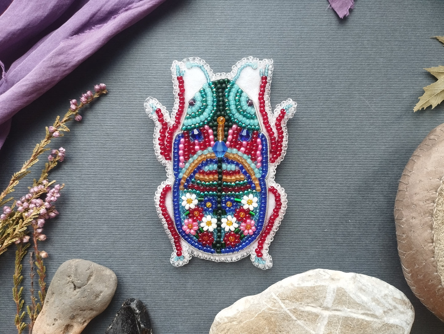 a colorful, intricately beaded object resembling a bug or insect, surrounded by various natural elements such as flowers, rocks, and leaves.