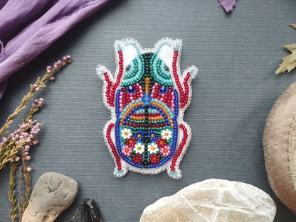 a colorful, intricately beaded object resembling a bug or insect, surrounded by various natural elements such as flowers, rocks, and leaves.
