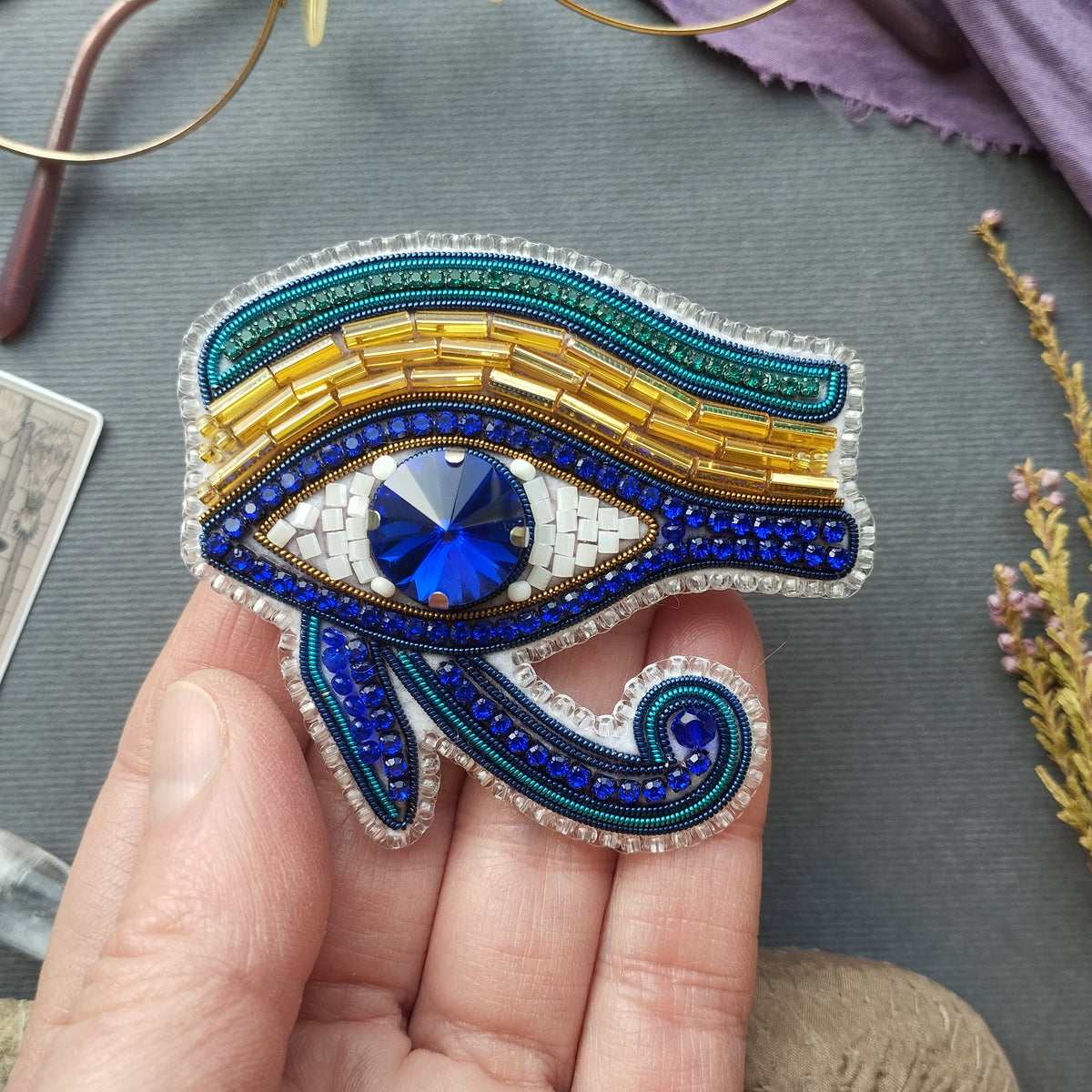 A hand holding a decorative eye brooch with a blue gemstone in the center.