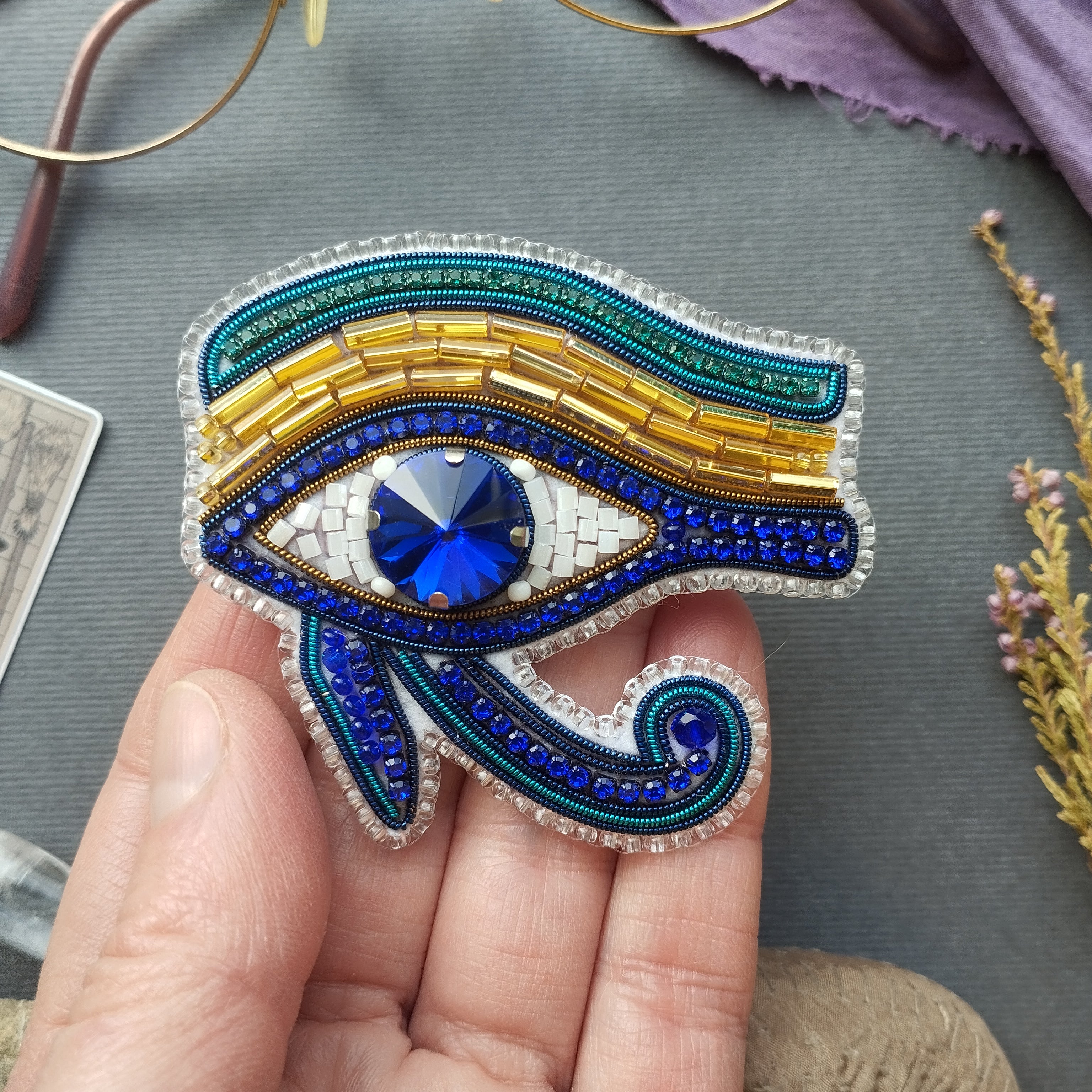 A hand holding a decorative eye brooch with a blue gemstone in the center.