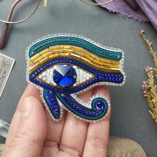 A hand holding a decorative eye brooch with a blue gemstone in the center.