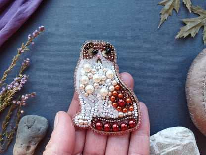 A hand holding a beaded cat-shaped object against a blue background, surrounded by various natural elements such as leaves and rocks.