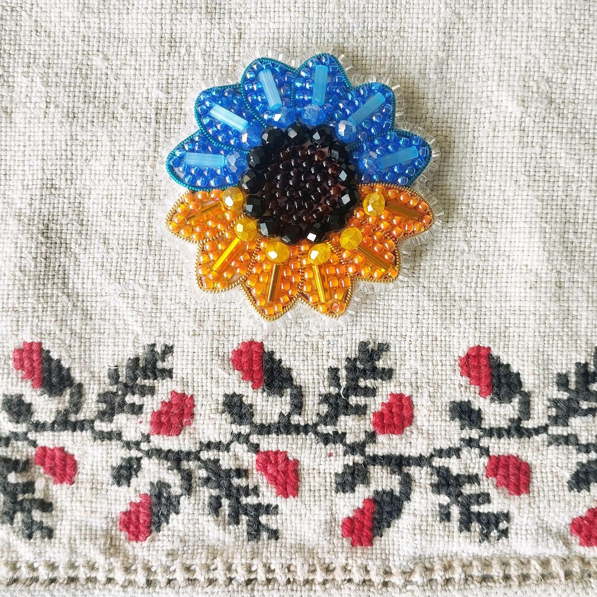 a close-up view of a decorative piece with a sunflower design, which is adorned with blue and orange beads. The piece is placed on a fabric with a floral pattern, creating a visually appealing contrast between the colorful beads and the intricate embroidery.