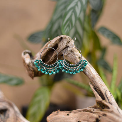 A pair of turquoise beaded earrings with circular hooks, displayed on a wooden branch against a blurred background of green leaves.