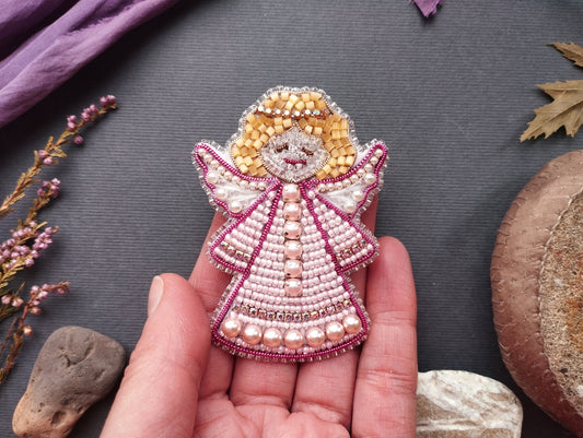 A hand holding a small, intricately beaded angel pin against a dark background.
