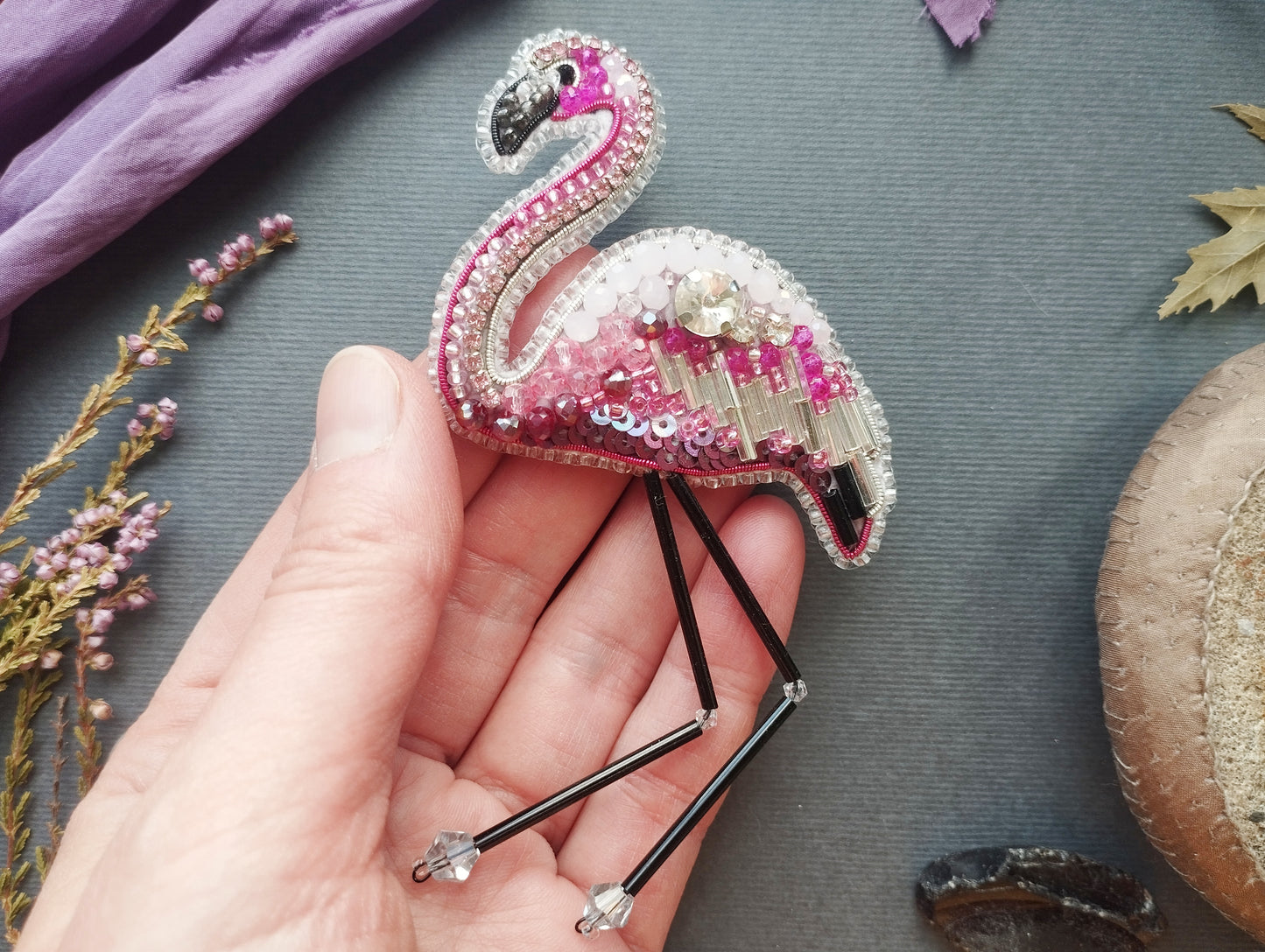 A hand holding a pink flamingo brooch with rhinestones and beads, surrounded by various objects such as leaves, flowers, and a mushroom.