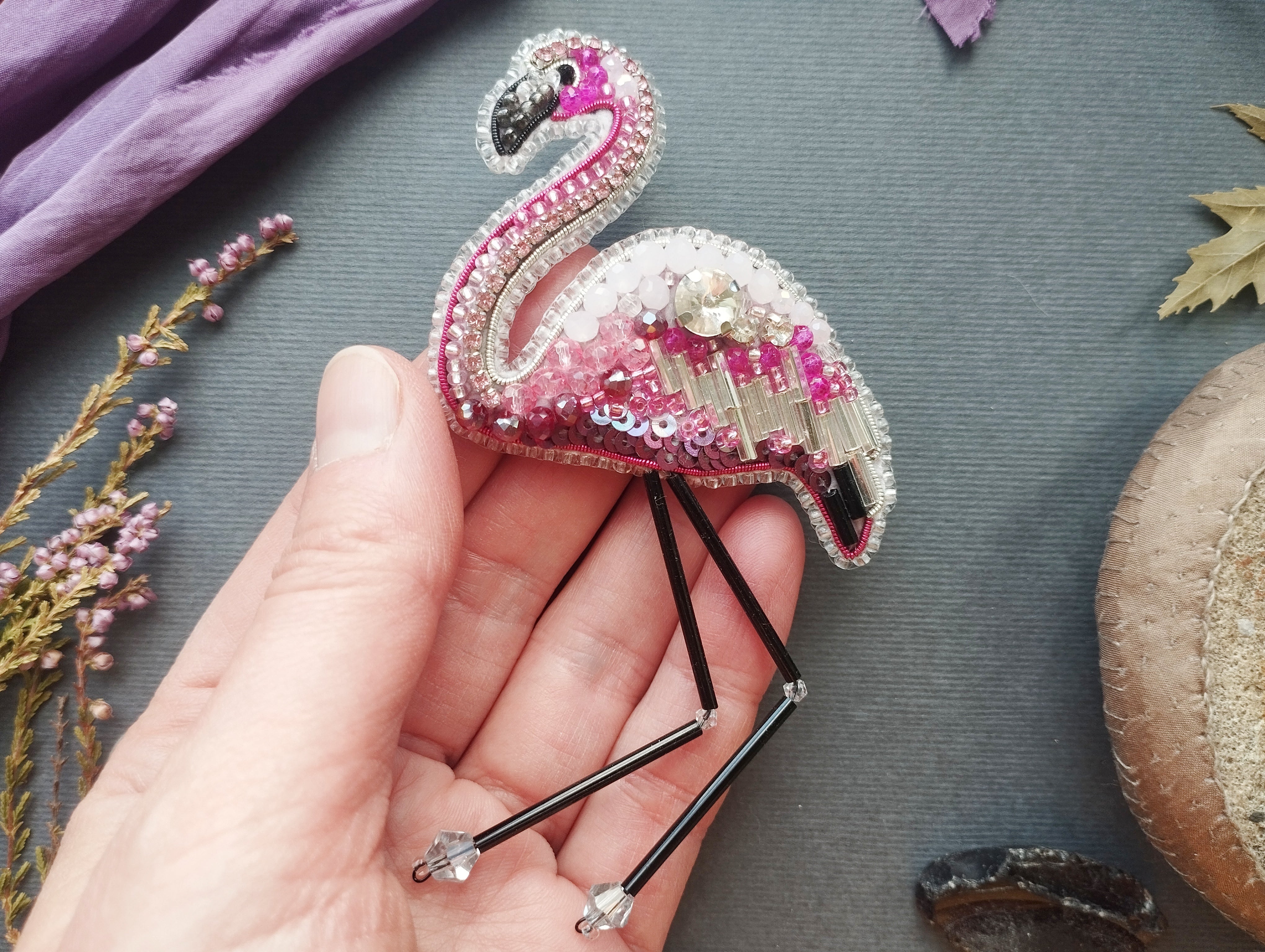 A hand holding a pink flamingo brooch with rhinestones and beads, surrounded by various objects such as leaves, flowers, and a mushroom.