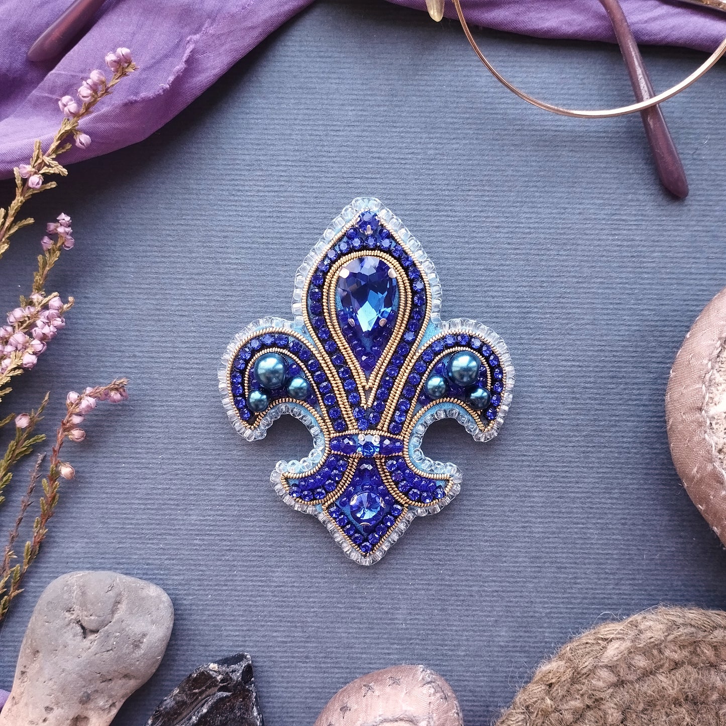 a blue and gold brooch with a large blue gemstone in the center, surrounded by smaller blue gemstones and a gold-colored gemstone. The brooch is placed on a gray surface, with some pink flowers and a pair of glasses visible in the background.