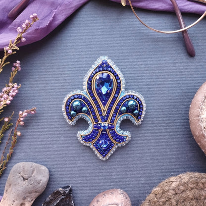 a blue and gold brooch with a large blue gemstone in the center, surrounded by smaller blue gemstones and a gold-colored gemstone. The brooch is placed on a gray surface, with some pink flowers and a pair of glasses visible in the background.