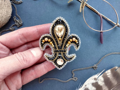 A hand holding a decorative brooch with a fleur-de-lis design, surrounded by various accessories such as glasses, a chain, and feathers.