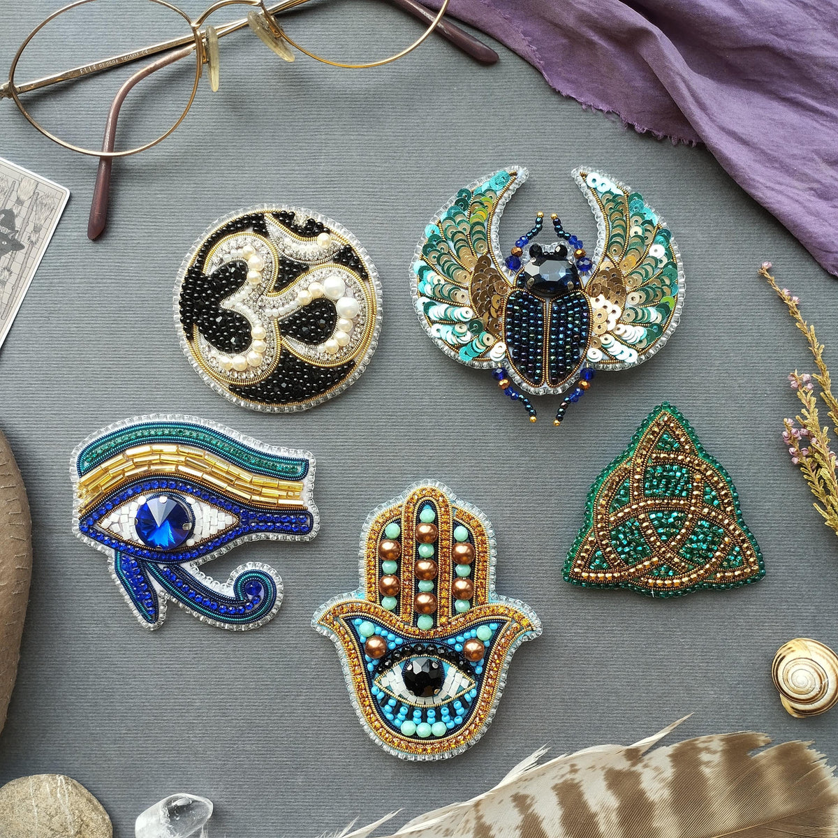 a collection of ornate, hand-made brooches and jewelry pieces, including a large eye-shaped brooch, a butterfly-shaped brooch, and a beetle-shaped brooch, all adorned with intricate beadwork and embellishments.