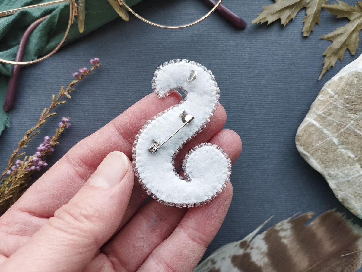 A hand holding a white fabric piece with a silver pin attached to it, surrounded by various objects such as a pair of glasses, leaves, and feathers.