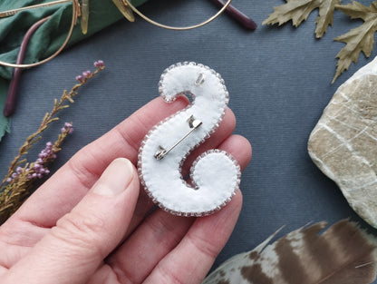 A hand holding a white fabric piece with a silver pin attached to it, surrounded by various objects such as a pair of glasses, leaves, and feathers.
