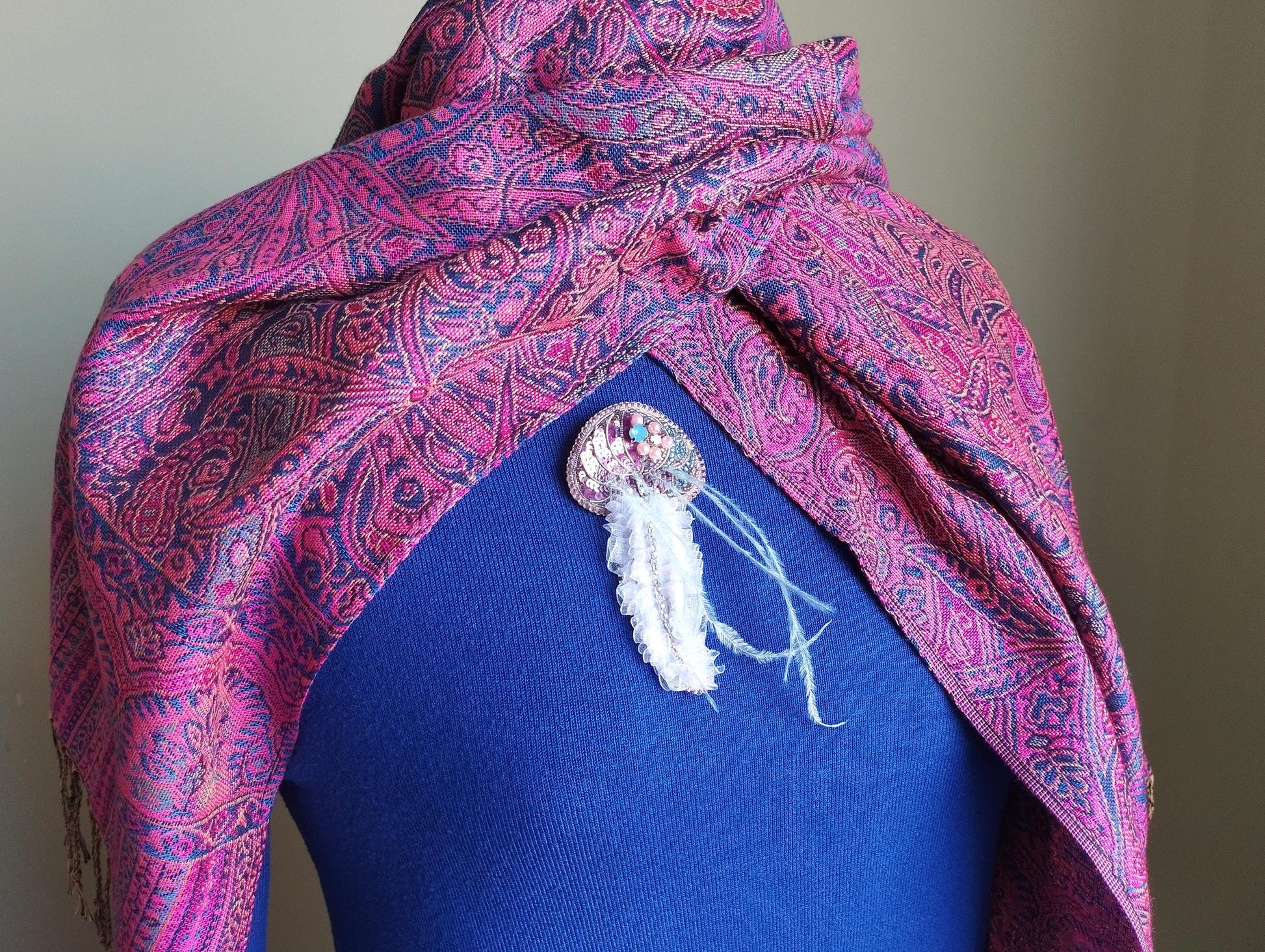 A mannequin wearing a blue top with a pink and purple paisley patterned shawl draped over it. The shawl has a white feather accessory hanging from it.