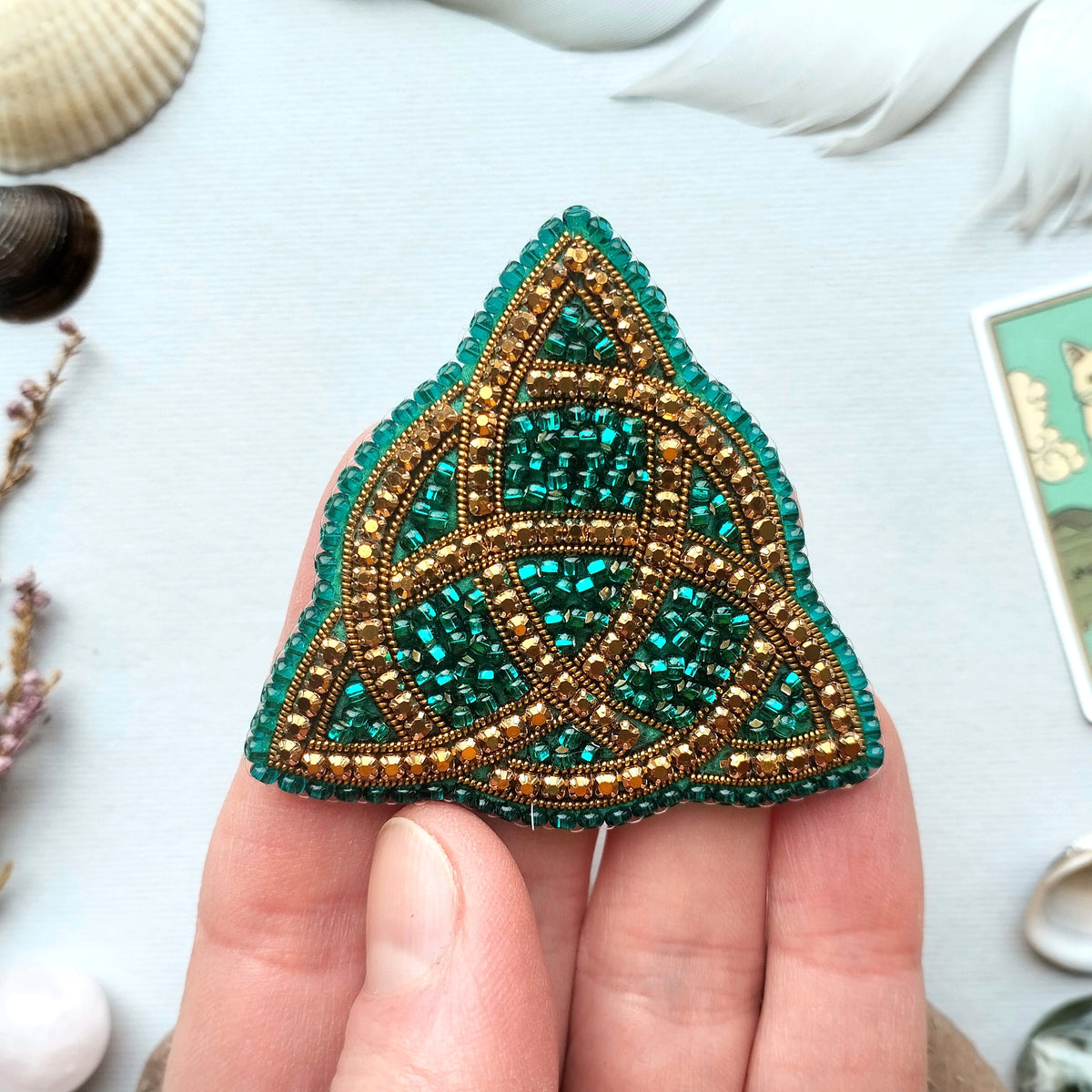A hand holding a triangular-shaped, intricately beaded object with a green and gold color scheme.
