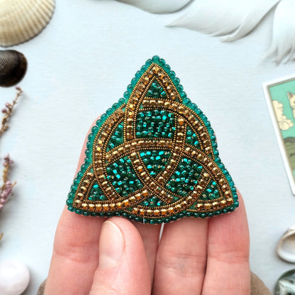 A hand holding a triangular-shaped, intricately beaded object with a green and gold color scheme.