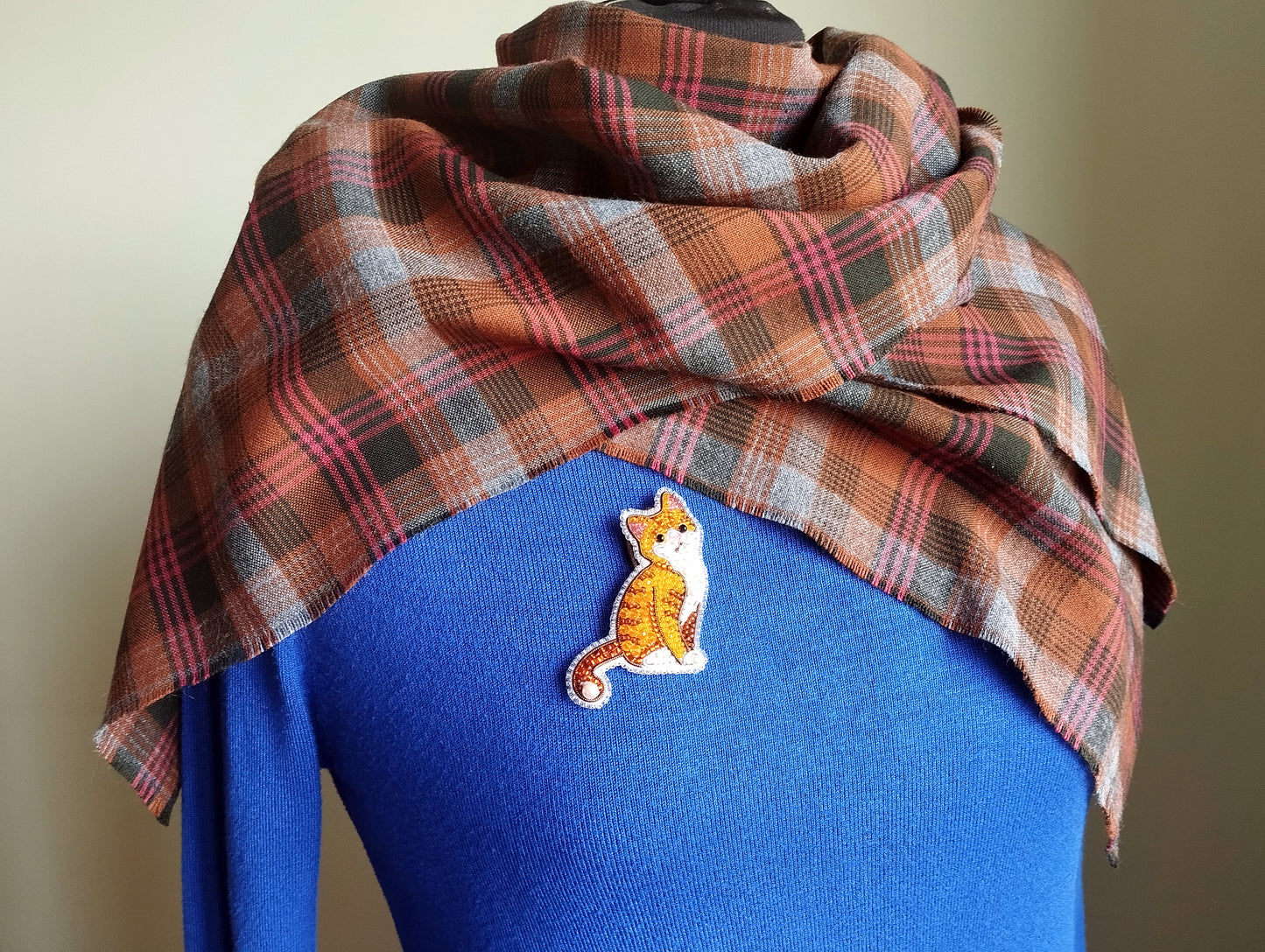 A plaid scarf is draped over a blue sweater, with a small embroidered cat pin visible on the sweater.