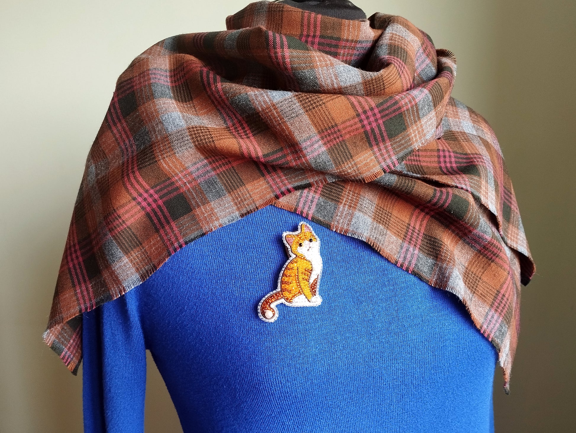 A plaid scarf is draped over a blue sweater, with a small embroidered cat pin visible on the sweater.