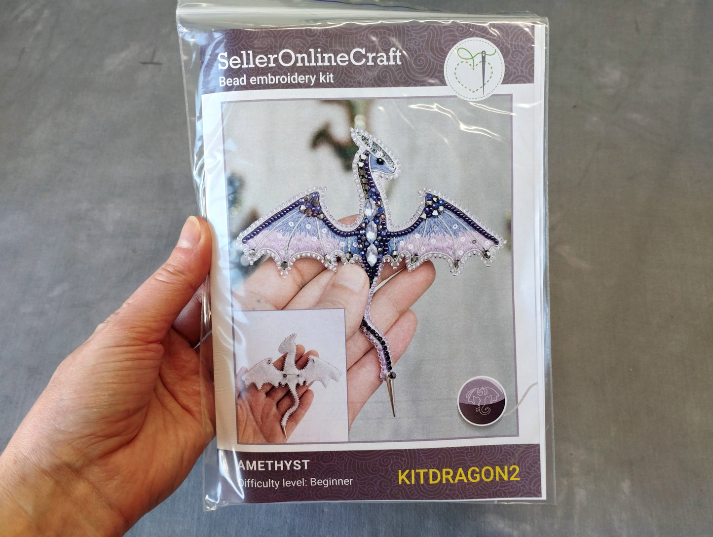 A hand holding a package containing a bead embroidery kit for creating a kit dragon.