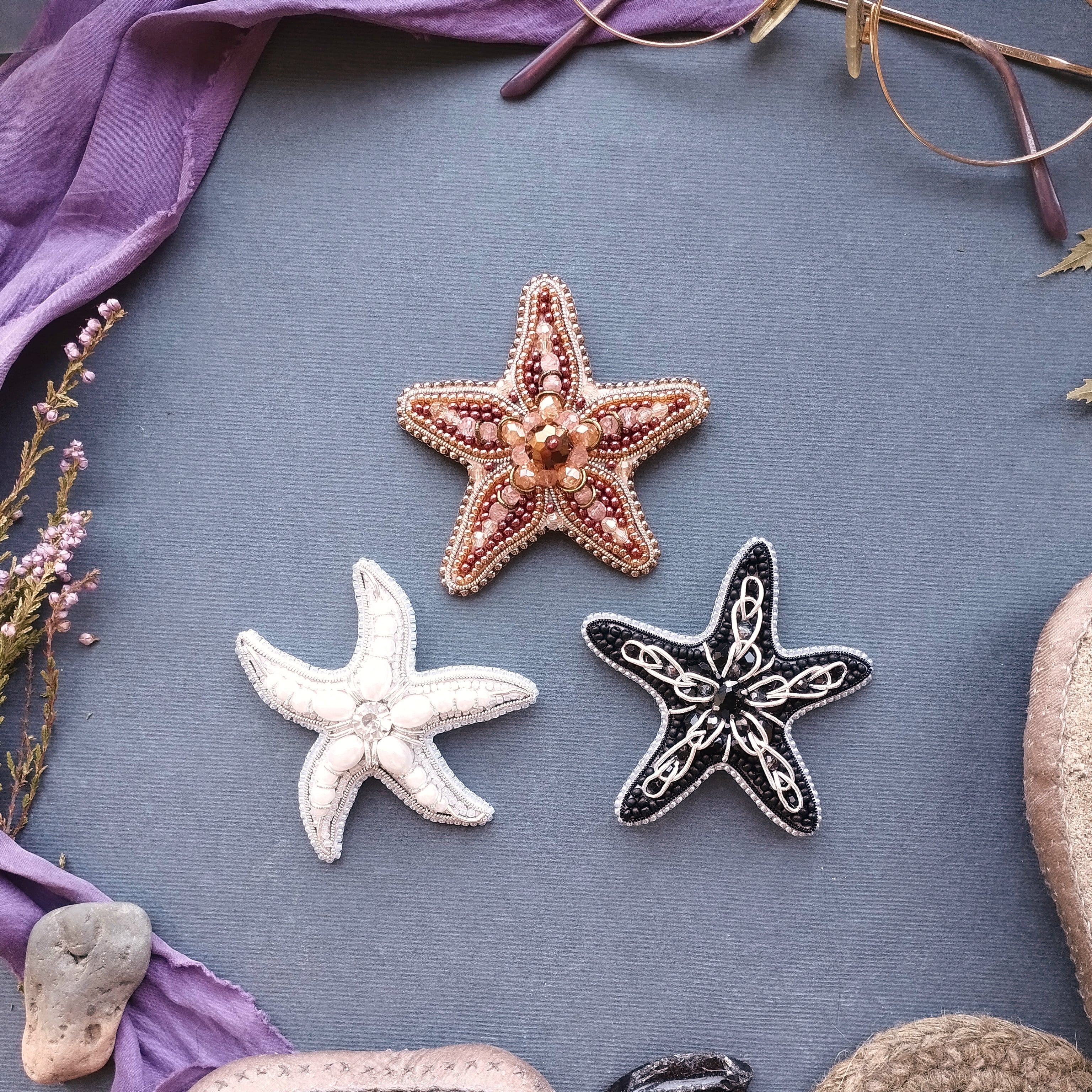 three starfish or sea starlets, each with a unique design, arranged on a blue background. The starfish on the left is white, the one in the middle is brown, and the one on the right is black. The starfish on the left appears to be the largest, while the black starfish on the right is the smallest. The starfish on the right is positioned slightly higher than the other two, creating a sense of depth in the image.