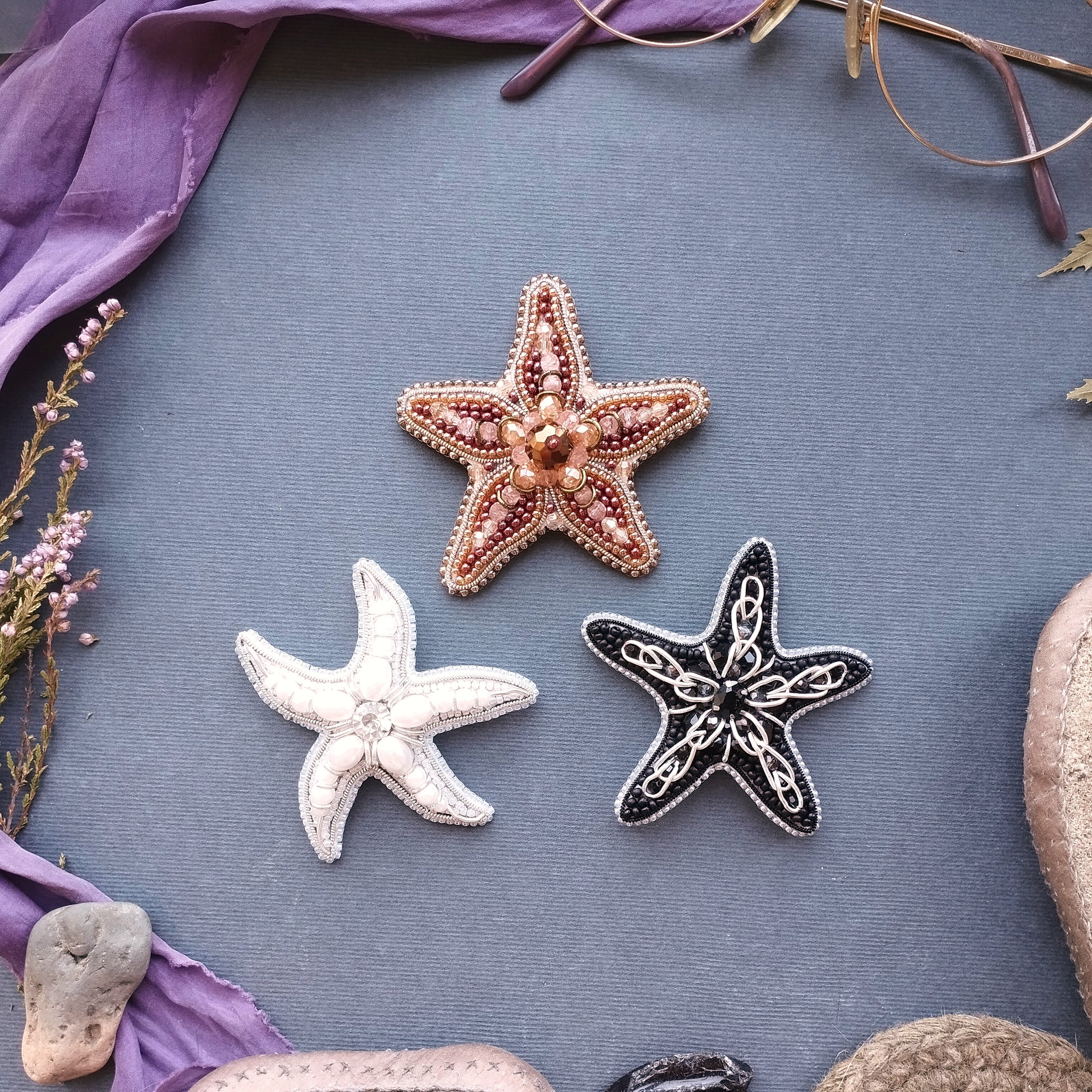 three starfish or sea starlets, each with a unique design, arranged on a blue background. The starfish on the left is white, the one in the middle is brown, and the one on the right is black. The starfish on the left appears to be the largest, while the black starfish on the right is the smallest. The starfish on the right is positioned slightly higher than the other two, creating a sense of depth in the image.