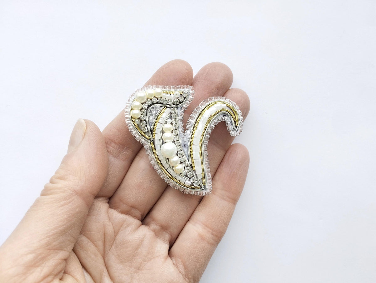 A hand holding a decorative piece of jewelry, which appears to be a brooch or pin, against a plain white background.