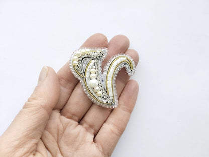 A hand holding a decorative piece of jewelry, which appears to be a brooch or pin, against a plain white background.