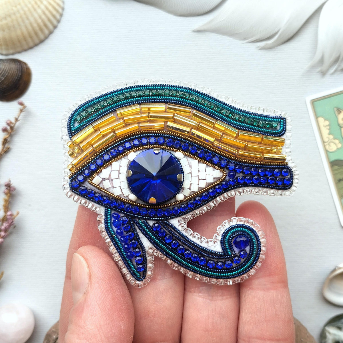 A hand holding a decorative eye brooch with a blue gemstone in the center, surrounded by various beads and shells.