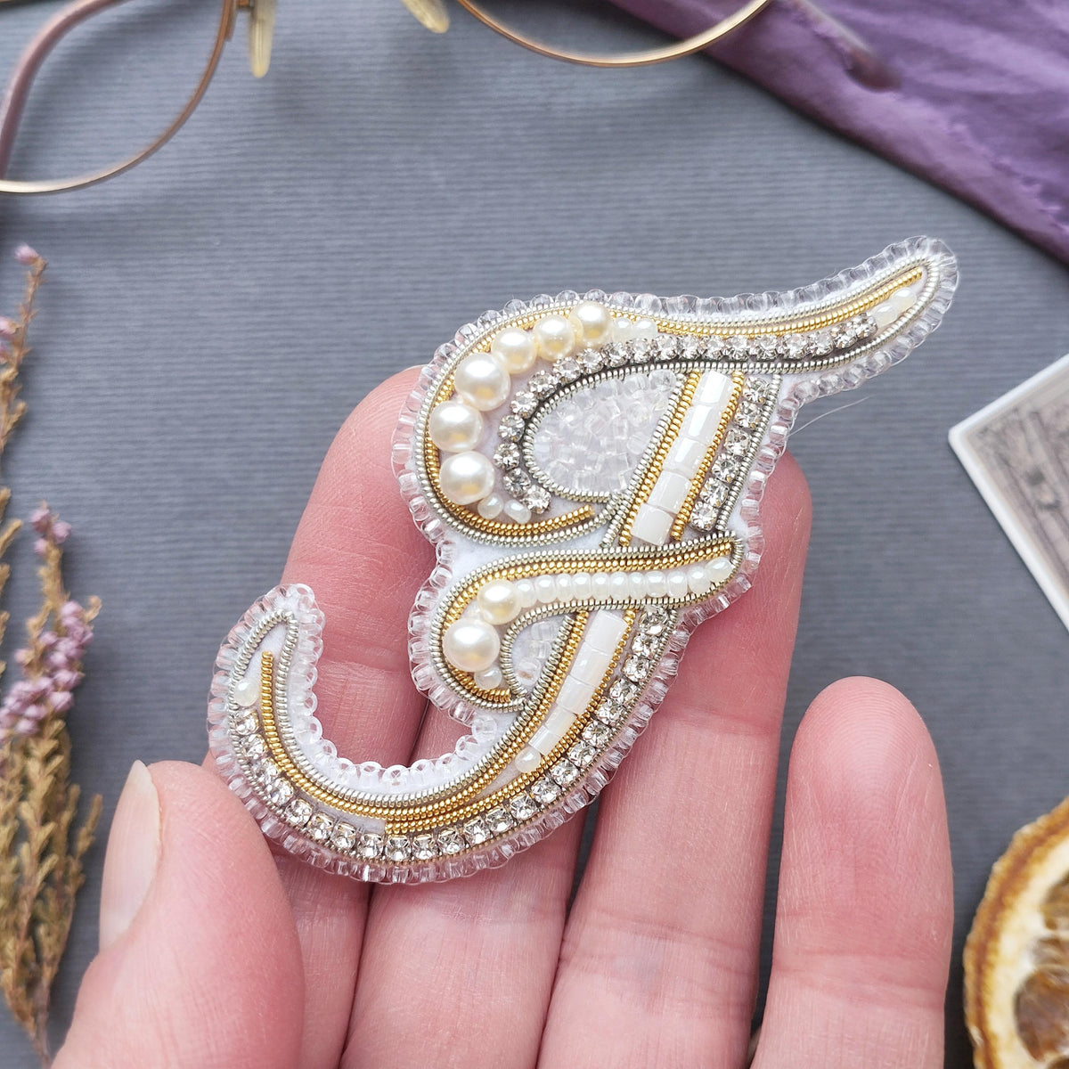 A hand holding a decorative letter 'F' made of beaded and embroidered materials, with a purple background and some dried flowers in the background.