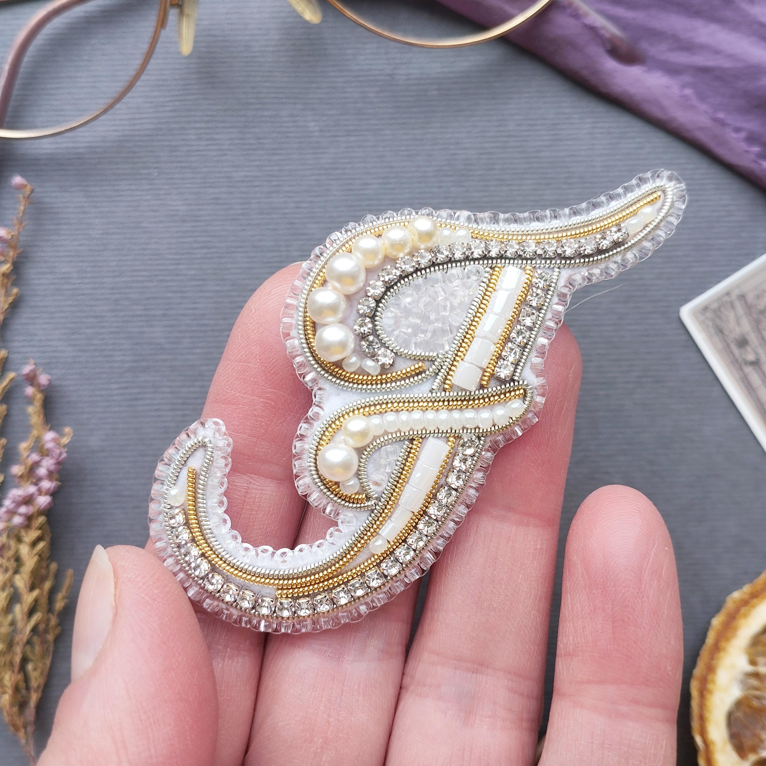 A hand holding a decorative letter 'F' made of beaded and embroidered materials, with a purple background and some dried flowers in the background.