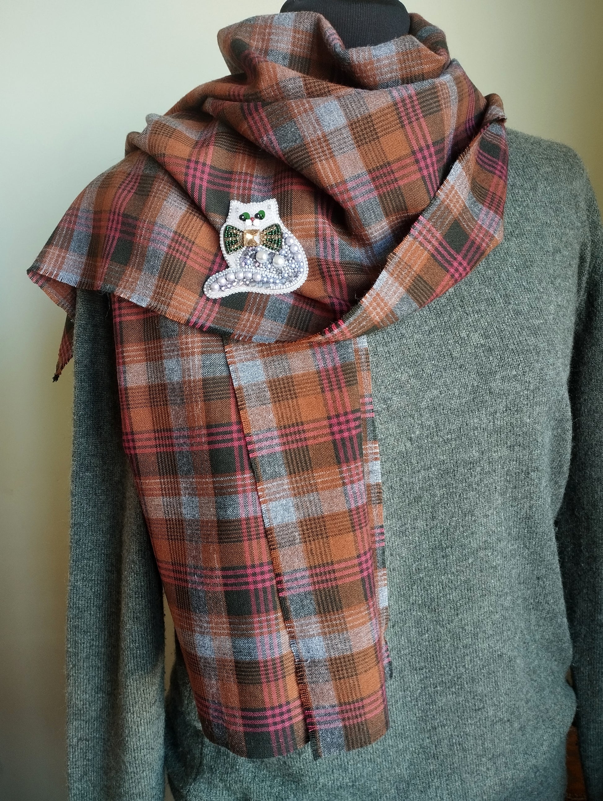A mannequin wearing a gray sweater and a plaid scarf with a small white owl pin on it.