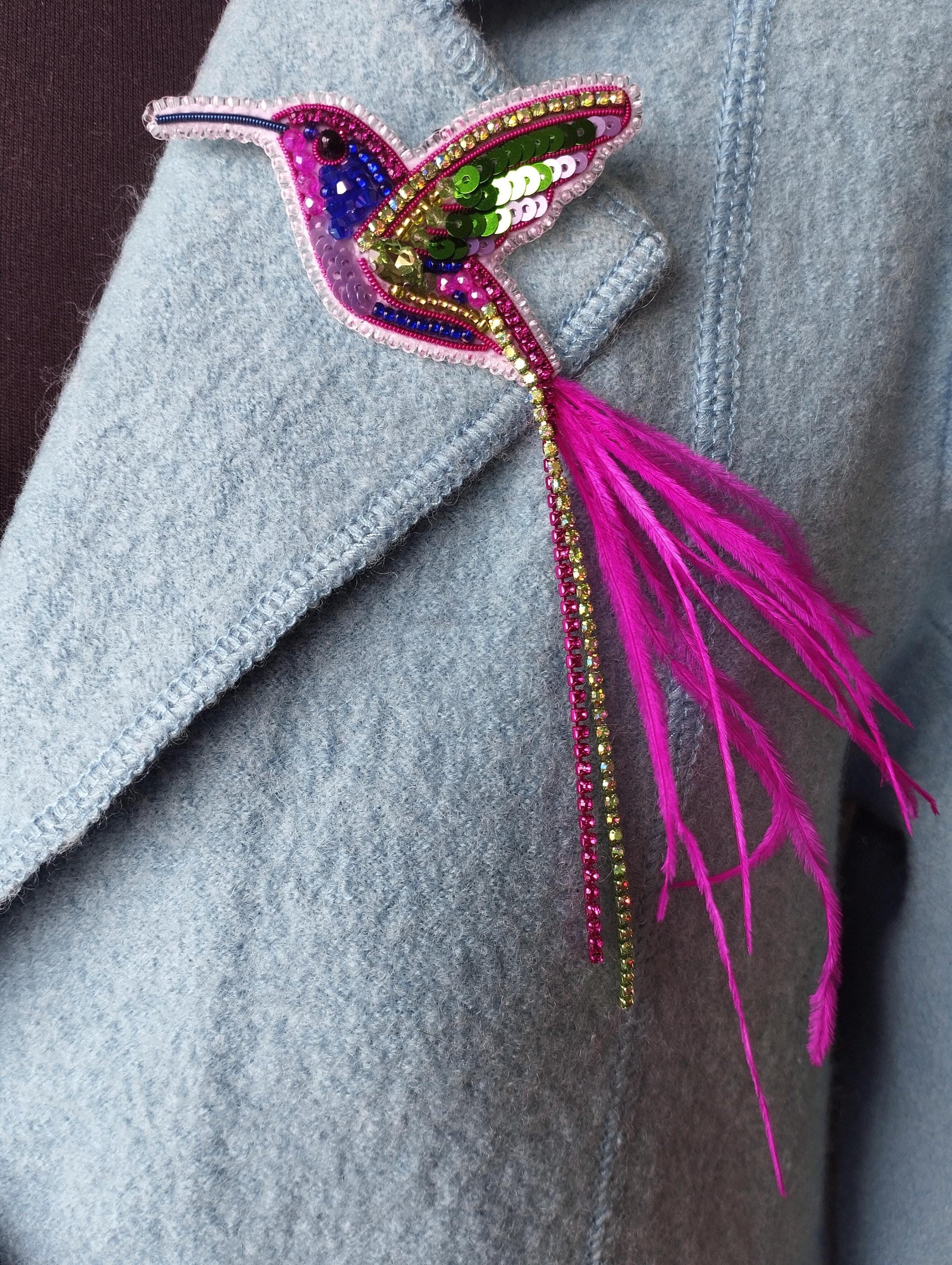 A close-up of a light blue jacket with a colorful, intricately designed hummingbird brooch or pin attached to the front.