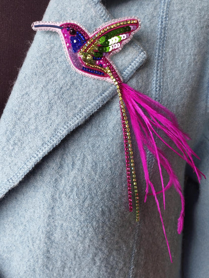 A close-up of a light blue jacket with a colorful, intricately designed hummingbird brooch or pin attached to the front.