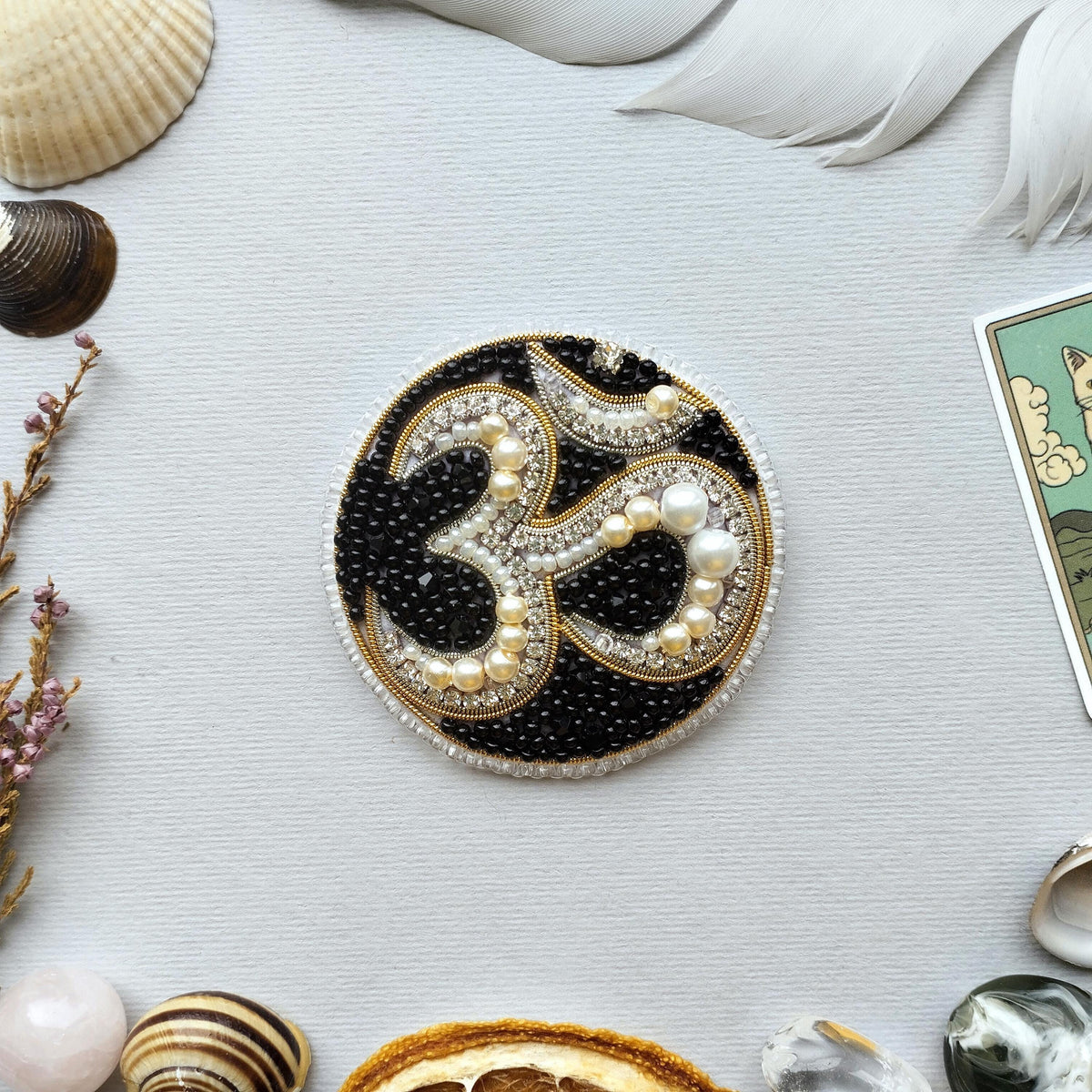 a circular piece of jewelry with a black and gold design, surrounded by various seashells, feathers, and other natural elements.