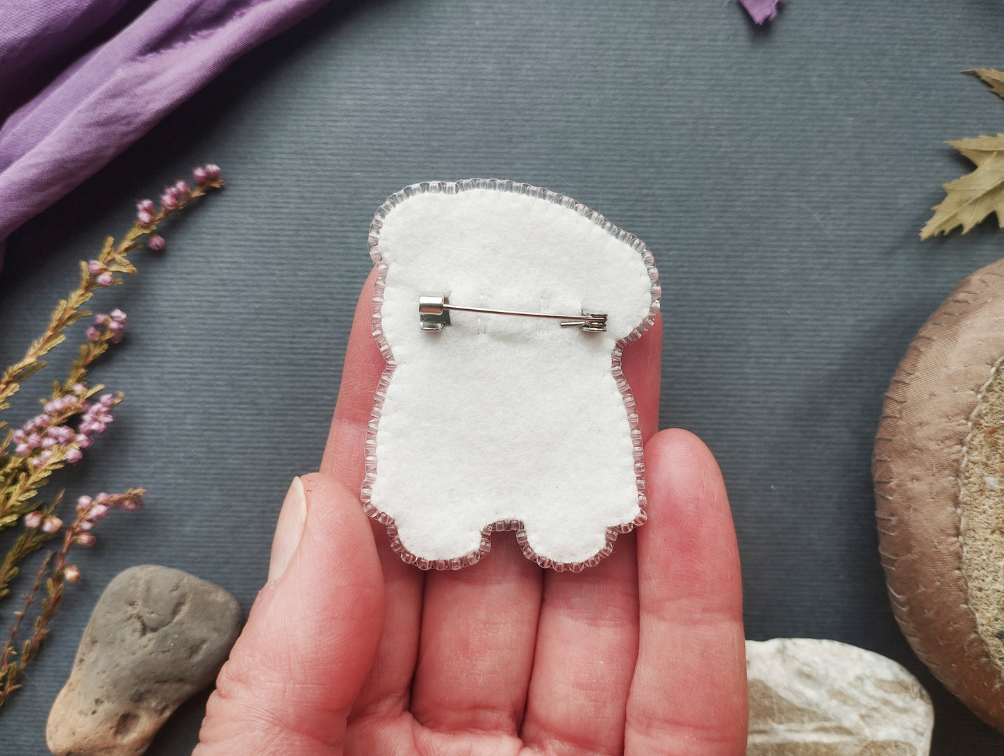 A hand holding a white felt object with a pin attached to it, surrounded by various natural elements such as flowers, rocks, and leaves.