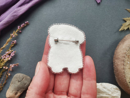 A hand holding a white felt object with a pin attached to it, surrounded by various natural elements such as flowers, rocks, and leaves.