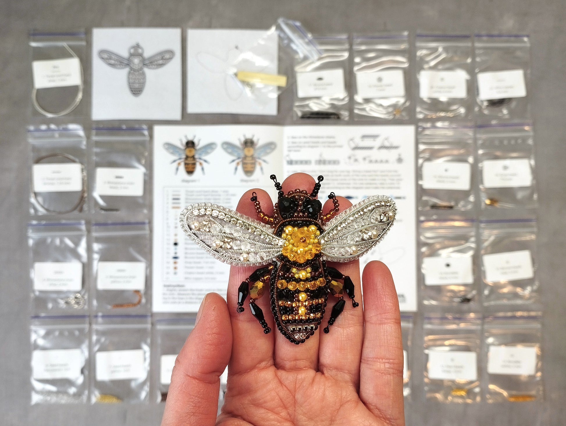 A hand is holding a decorative beehive-shaped brooch, which is made of silver and gold materials. The background includes various small packages and bags, possibly containing additional brooches or jewelry items.