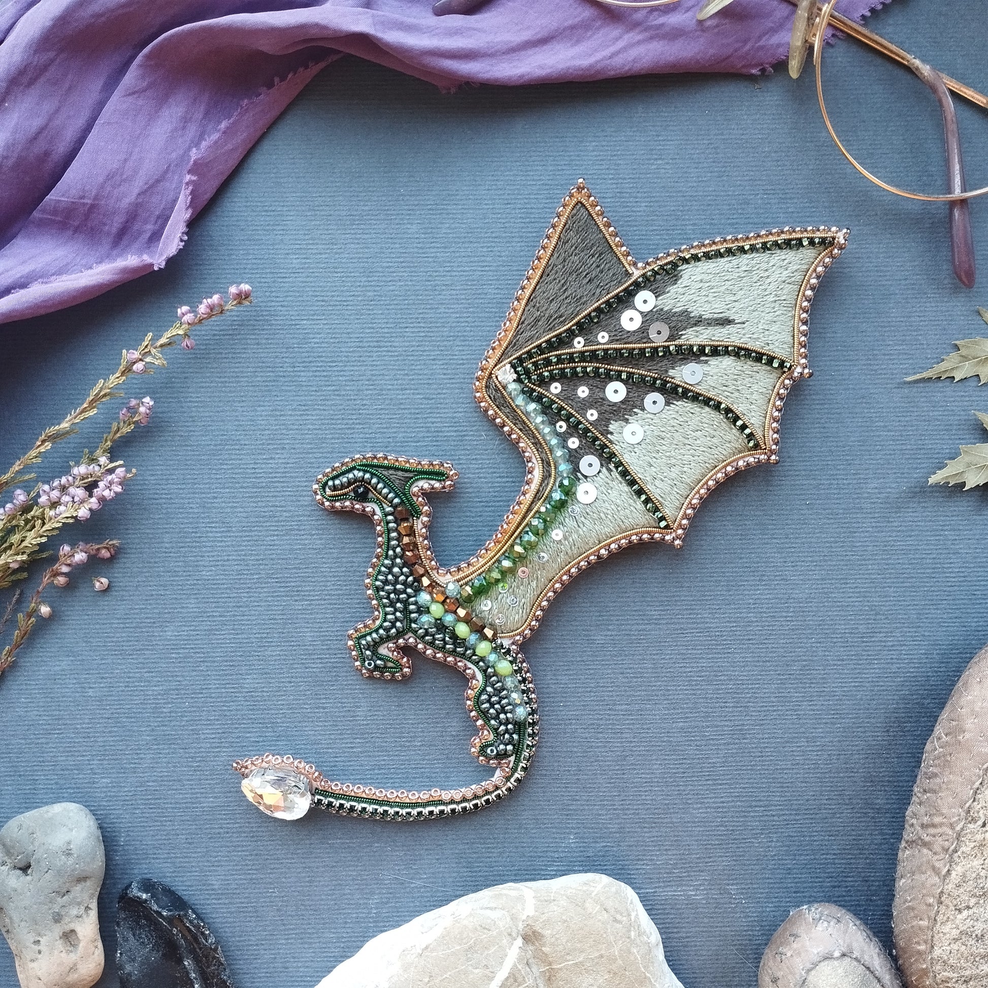 a dragon-shaped embroidered piece with a metallic and beaded design, placed on a blue surface surrounded by various natural elements such as rocks, flowers, and leaves.