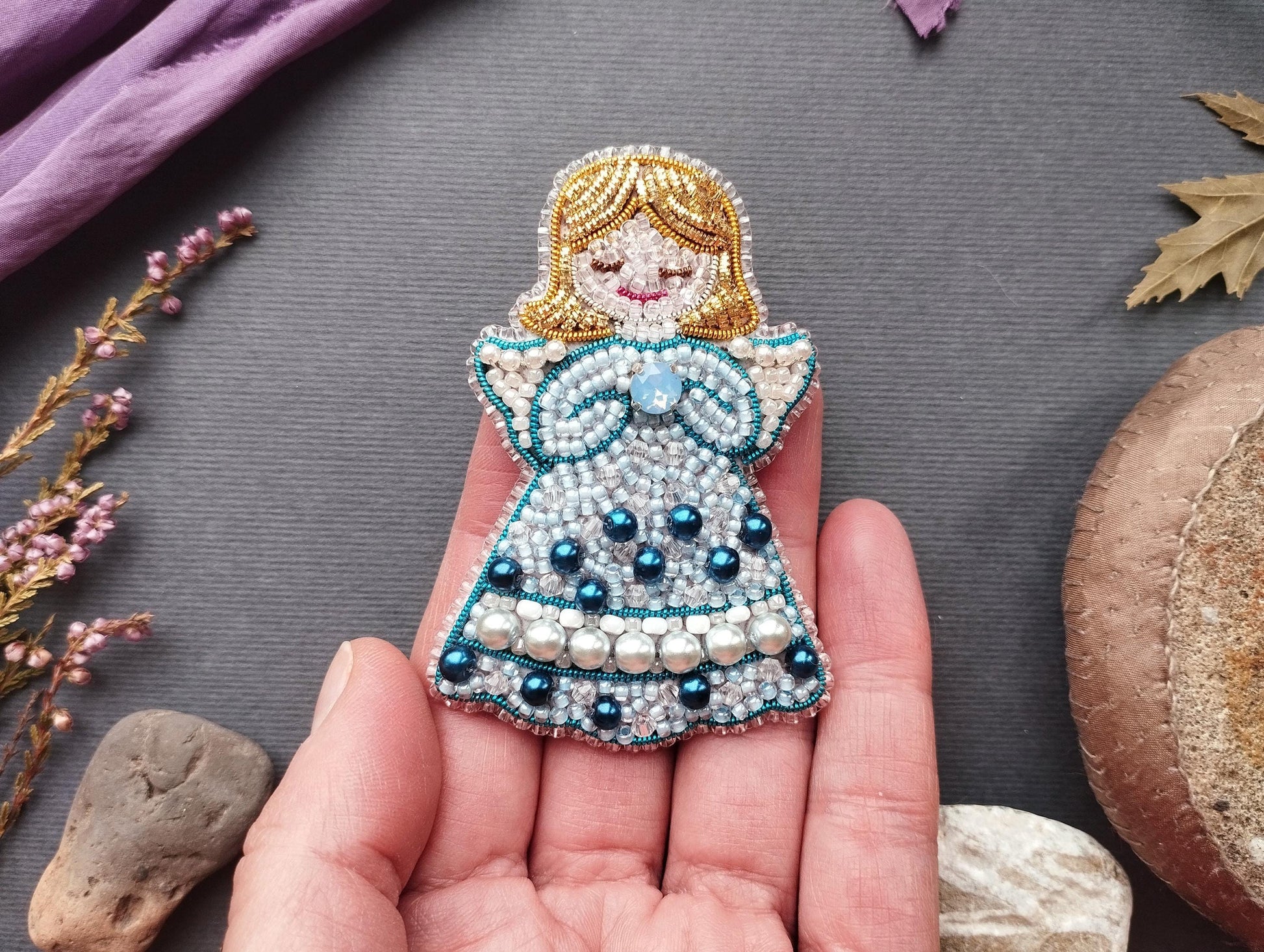 A hand holding a decorative beaded doll with a blue dress and pearl accents, surrounded by various natural elements such as flowers, leaves, and rocks.