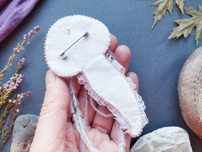 A hand holding a white, hand-sewn pin or badge with a delicate lace-like trim, surrounded by various natural elements such as leaves, flowers, and rocks.