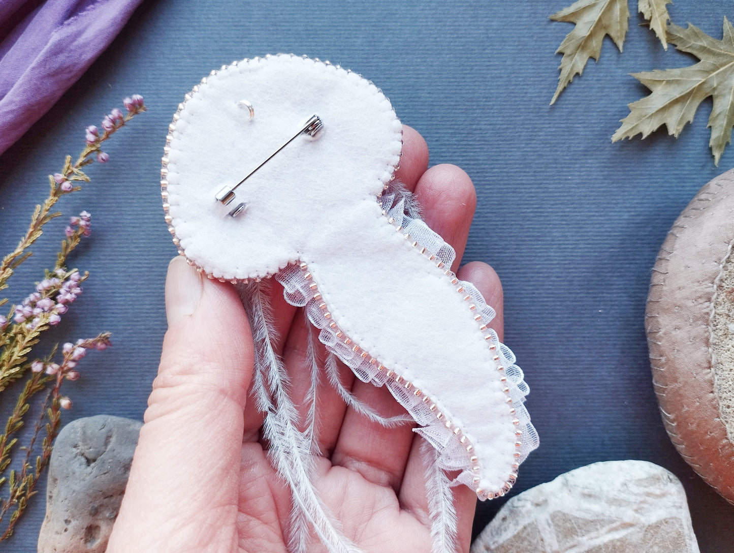 A hand holding a white, hand-sewn pin or badge with a delicate lace-like trim, surrounded by various natural elements such as leaves, flowers, and rocks.
