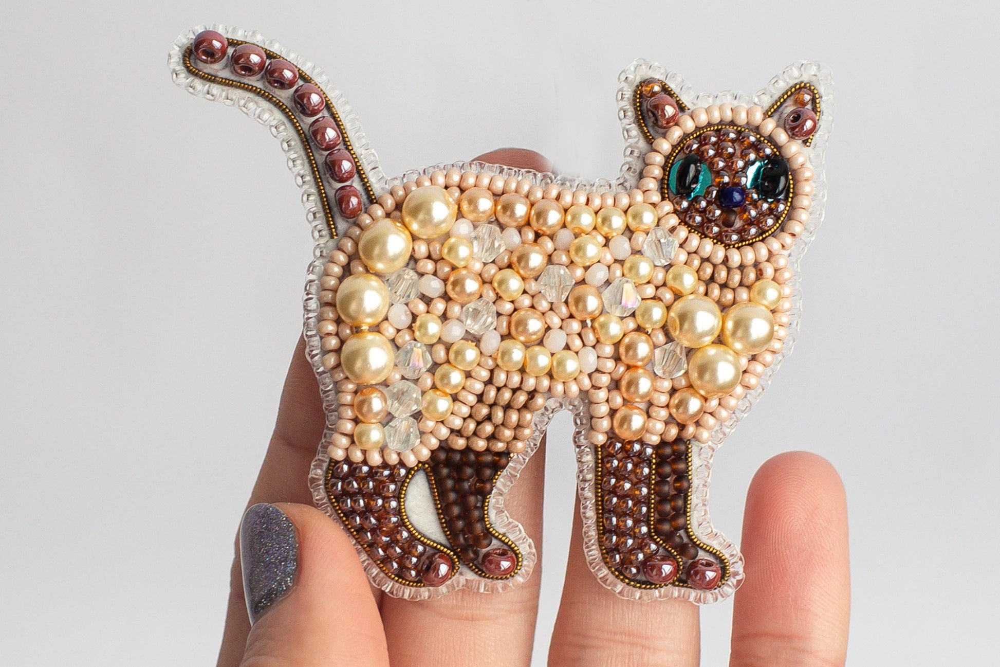 A hand holding a decorative cat pin made of beads and pearls.