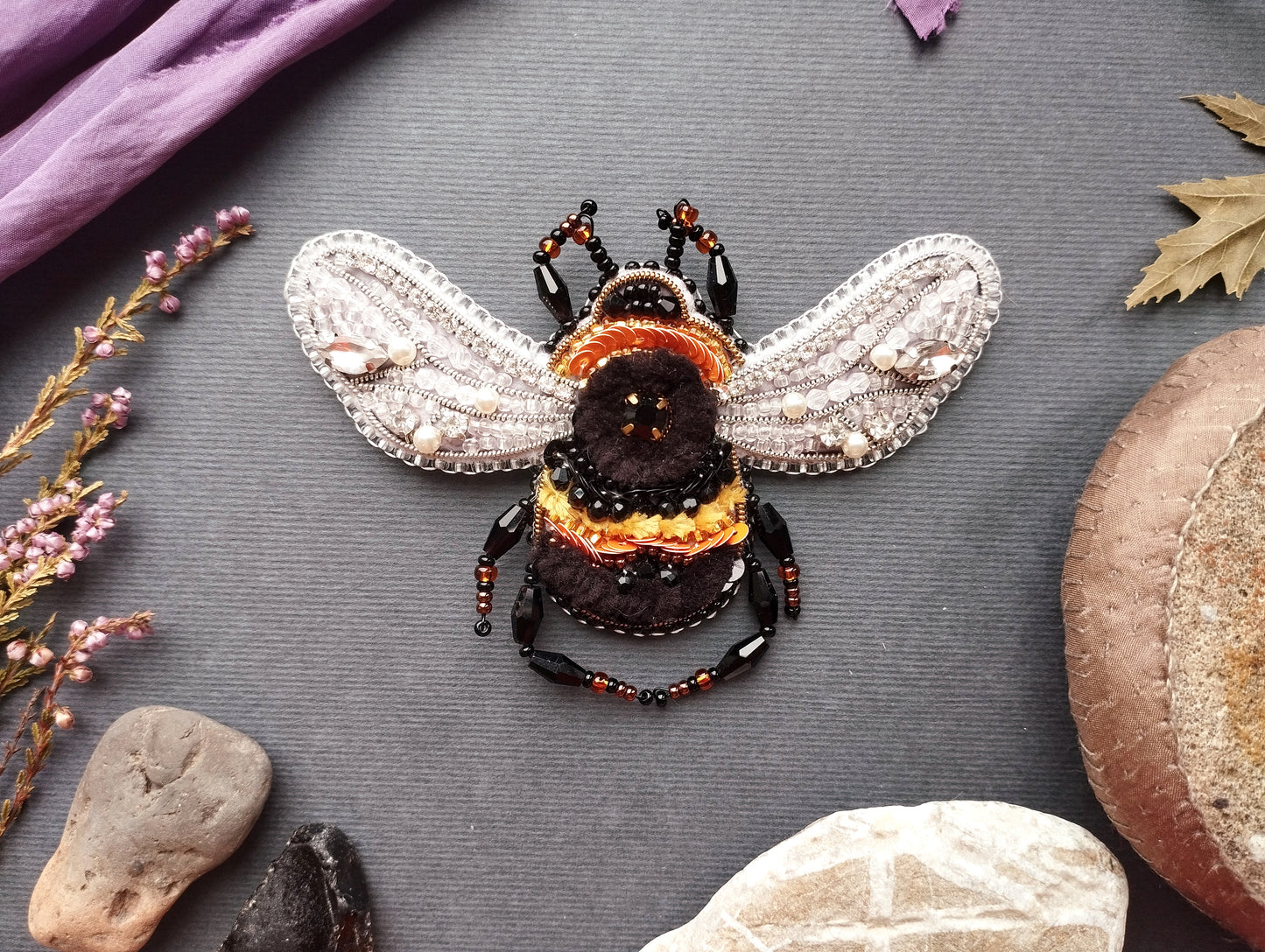 a detailed embroidered bee with intricate beading and beadwork, surrounded by various natural elements such as leaves, rocks, and flowers.