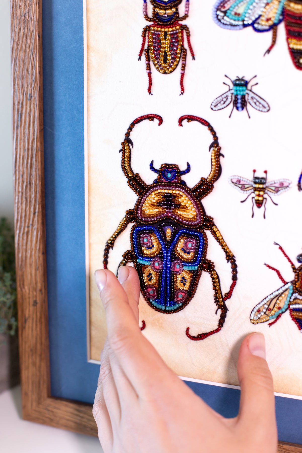 A hand is holding up a framed collage of colorful, intricately designed insects, including a large, detailed dragonfly.
