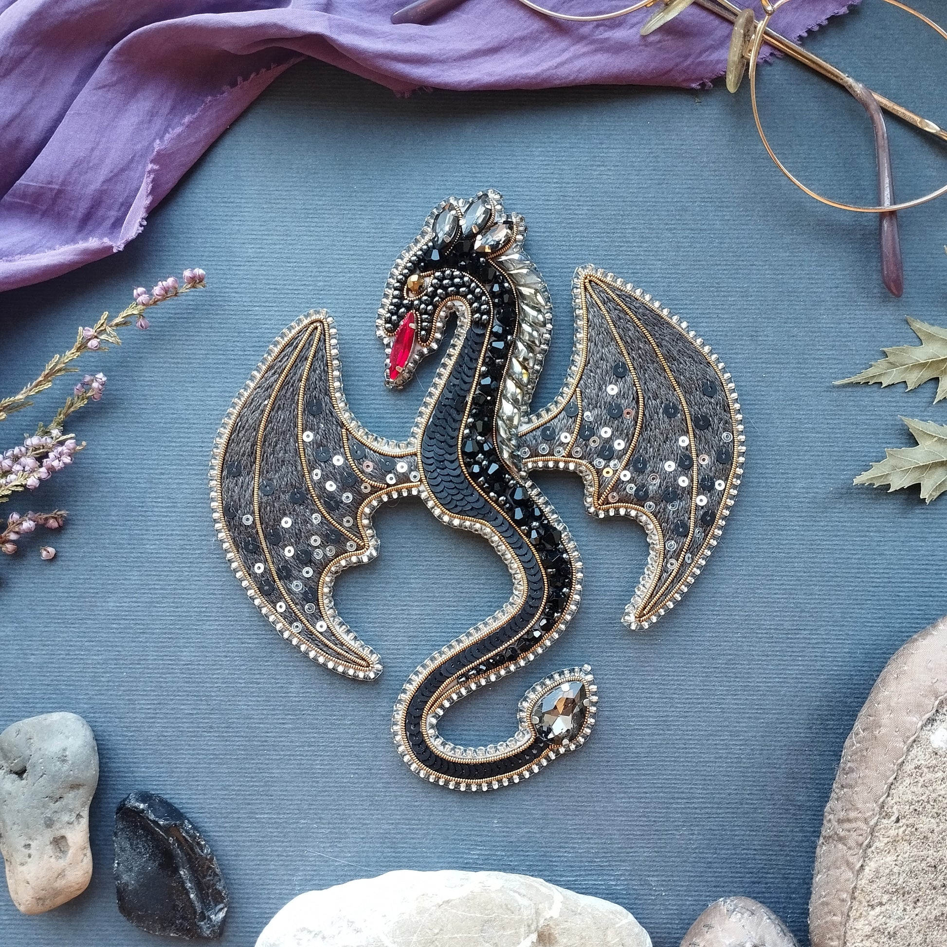 a detailed embroidered dragon brooch with intricate patterns and sparkling gemstones, surrounded by various natural elements such as leaves, rocks, and a purple fabric.
