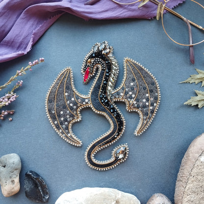 a detailed embroidered dragon brooch with intricate patterns and sparkling gemstones, surrounded by various natural elements such as leaves, rocks, and a purple fabric.
