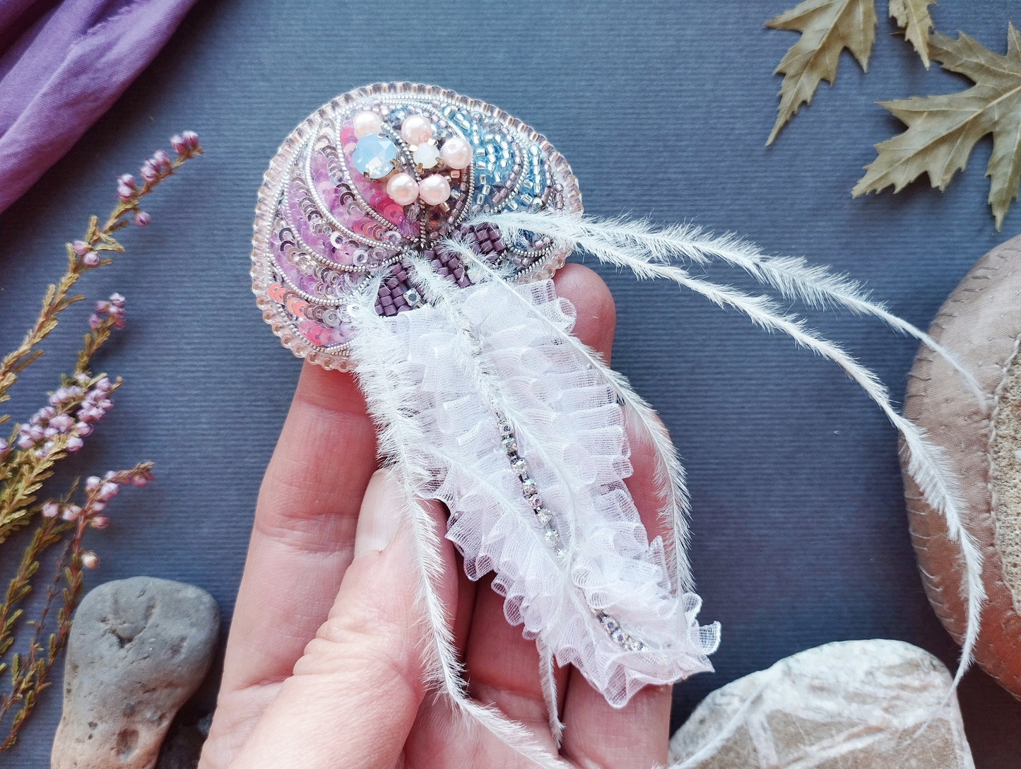 A hand holding a decorative piece with a ruffled white feather attached to it, surrounded by various natural elements such as leaves, flowers, and rocks.