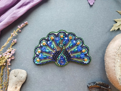 a decorative peacock feather-shaped brooch with intricate beading and gemstone details, placed on a gray background alongside various natural elements such as leaves, flowers, and rocks.