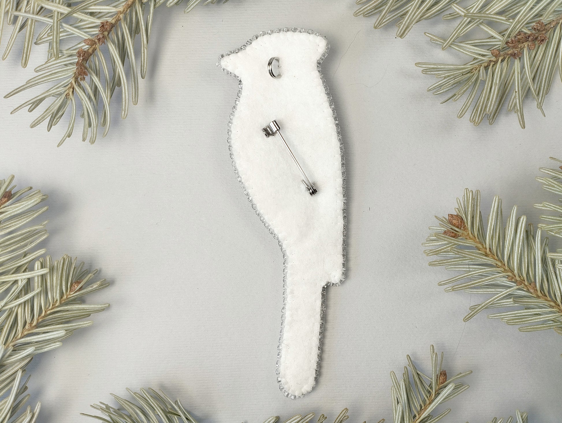 A white bird-shaped ornament with a silver pin attached to it, surrounded by pine needles on a light gray background.