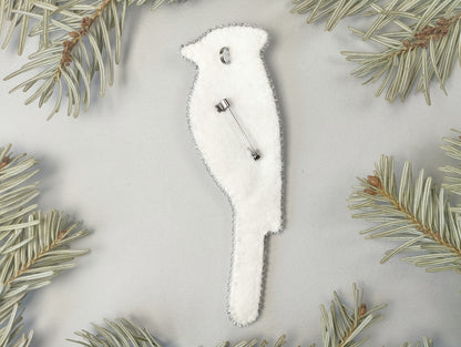 A white bird-shaped ornament with a silver pin attached to it, surrounded by pine needles on a light gray background.
